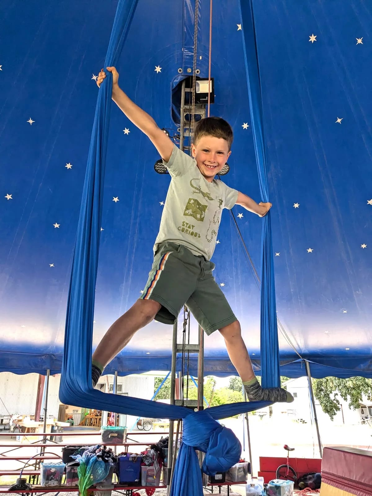 A grinning camper stands on aerial silks knotted below him, arms outstretched, inside the school's star-patterned blue circus tent