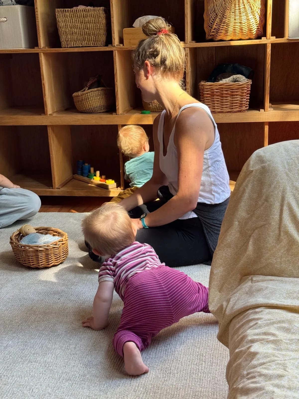 A teacher stays low on the rug in the toddler room as a baby crawls over to her.