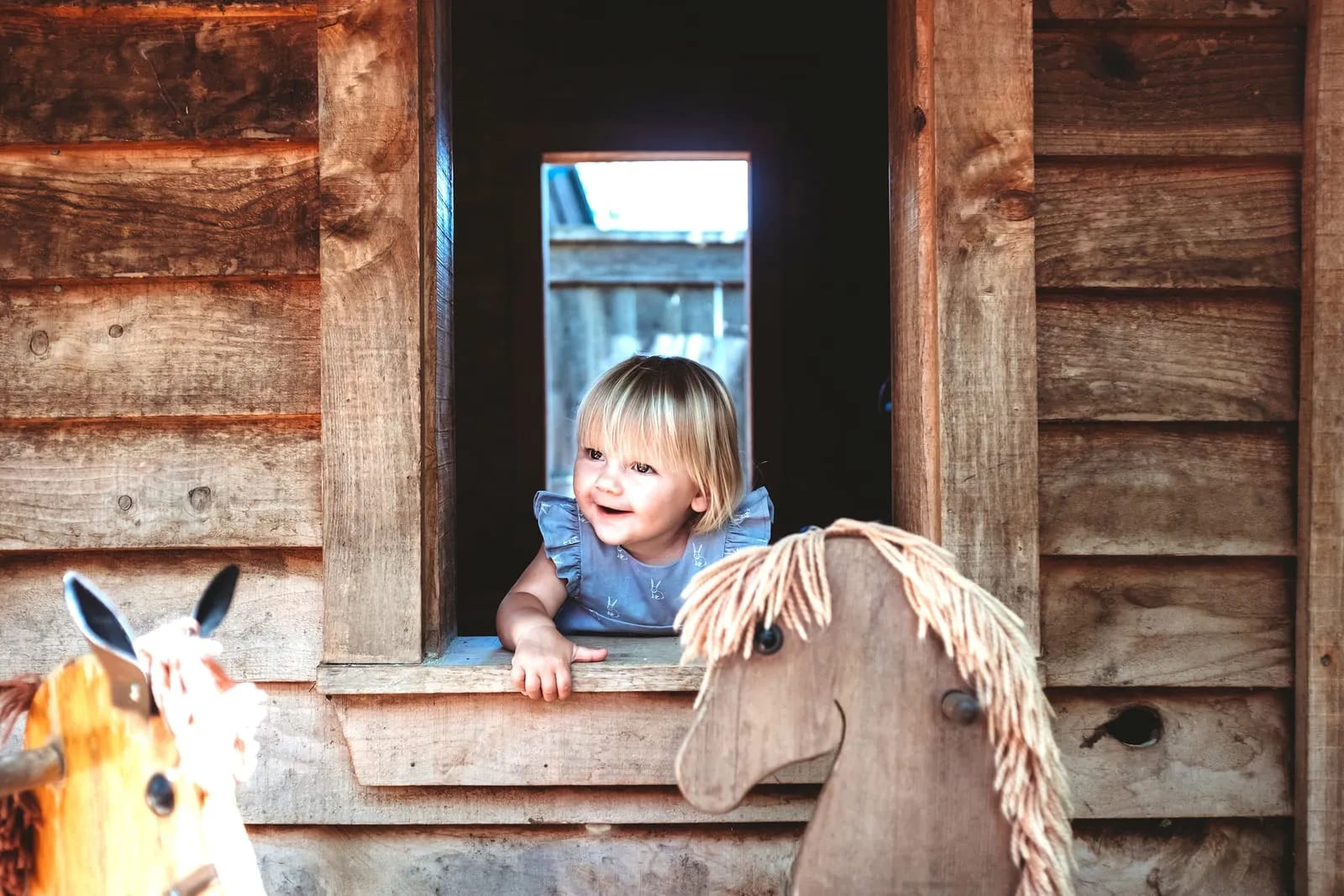 A child leans out of the playhouse window, wooden hobby horses parked below.